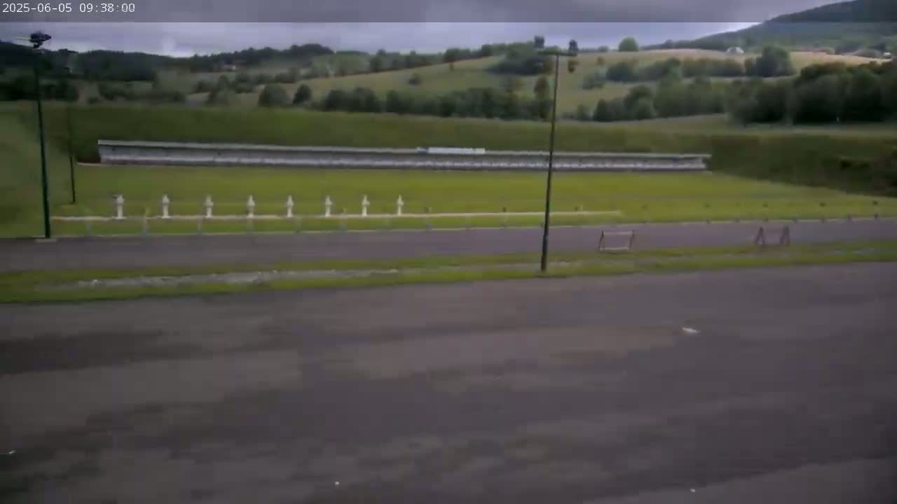 An outdoor shooting range with a long, low wall, several targets, and a paved area sits under an overcast sky.