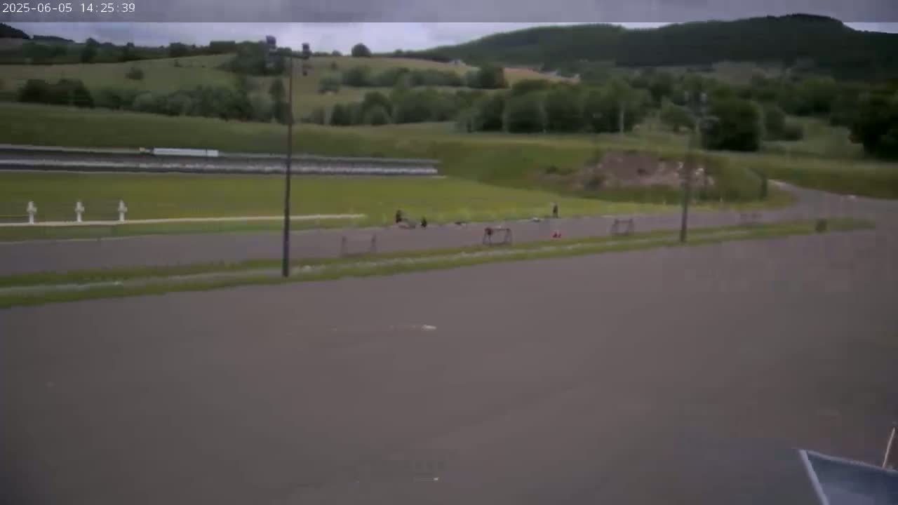 A wide shot of an overcast day shows a paved area with several people scattered about, near a grassy area with a backdrop of rolling hills and trees.