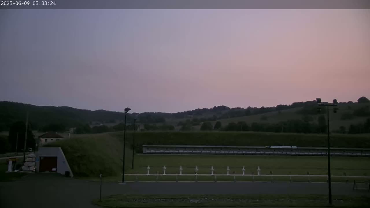 An outdoor shooting range is situated on a grassy field, backed by rolling hills under a dusky pink and purple sky.