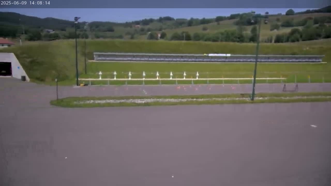 A long, gray asphalt area curves in front of a shooting range set against a grassy hillside under an overcast sky.