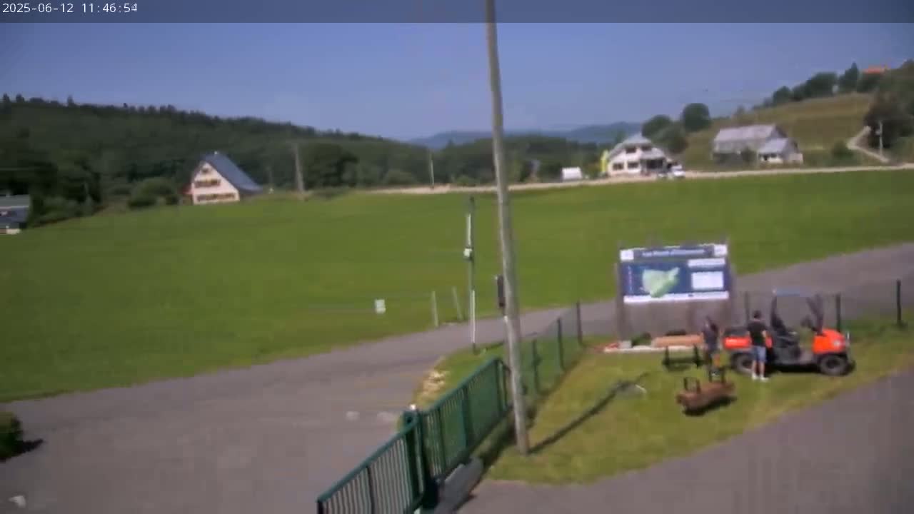A sunny day shows a grassy field with a few buildings and trees in the background, an orange utility vehicle, and a few people near a gate and information board.