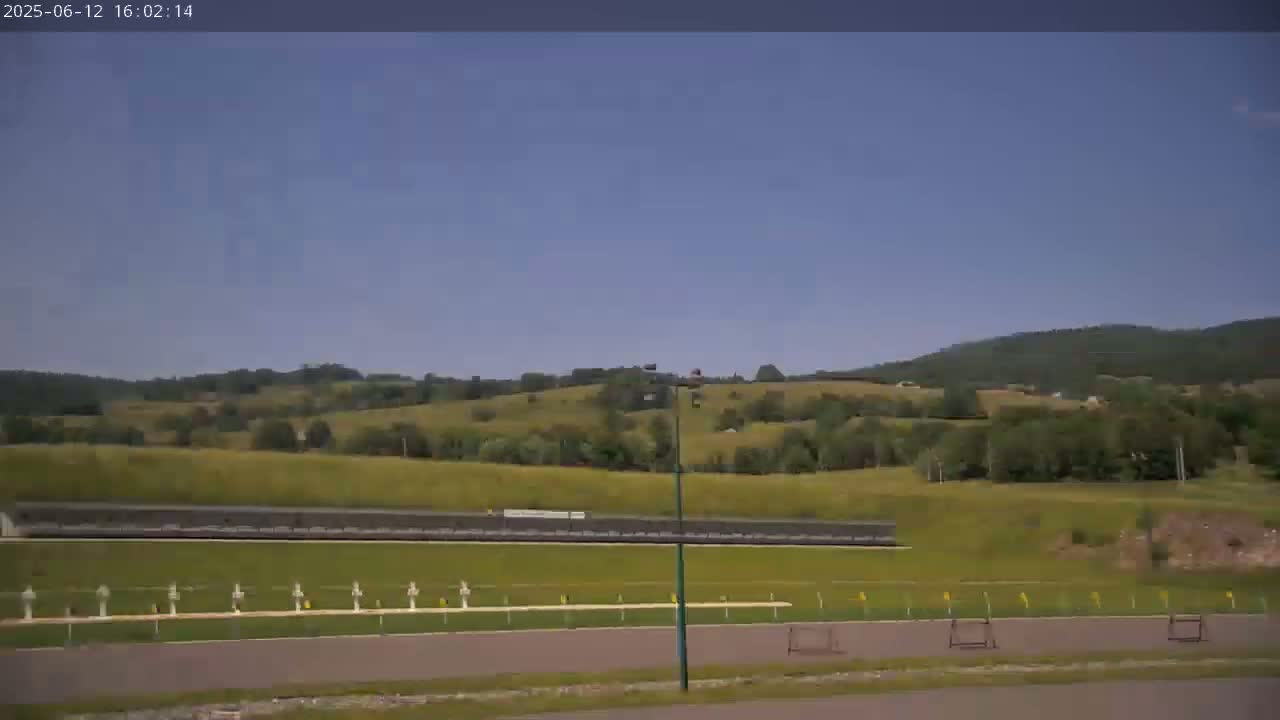 A shooting range is situated in a valley, with rolling green hills and a clear blue sky in the background.