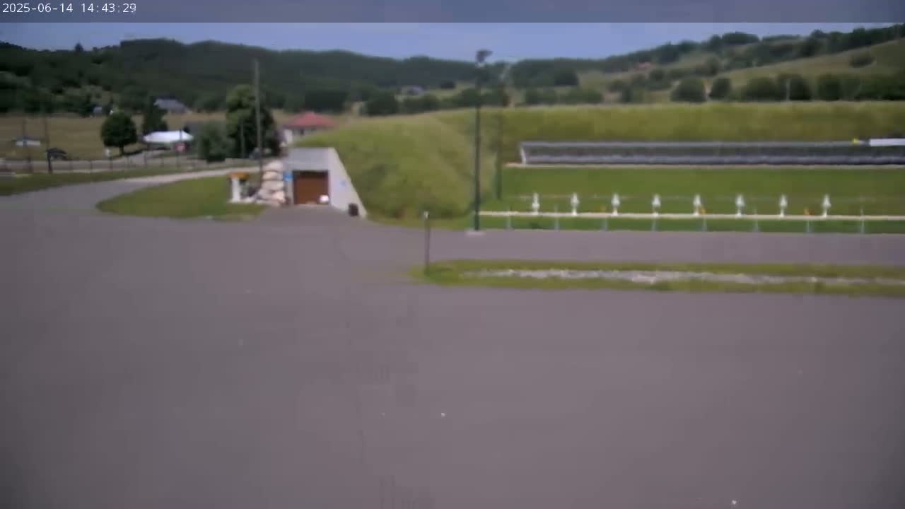 A paved area curves toward a building with a garage door, beyond which is a grassy area with a row of white markers and rolling green hills under an overcast sky.