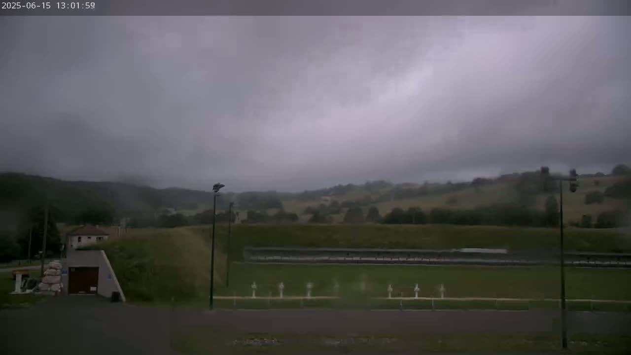 An overcast sky hangs over a grassy field with a shooting range, a small building, and rolling hills in the background.