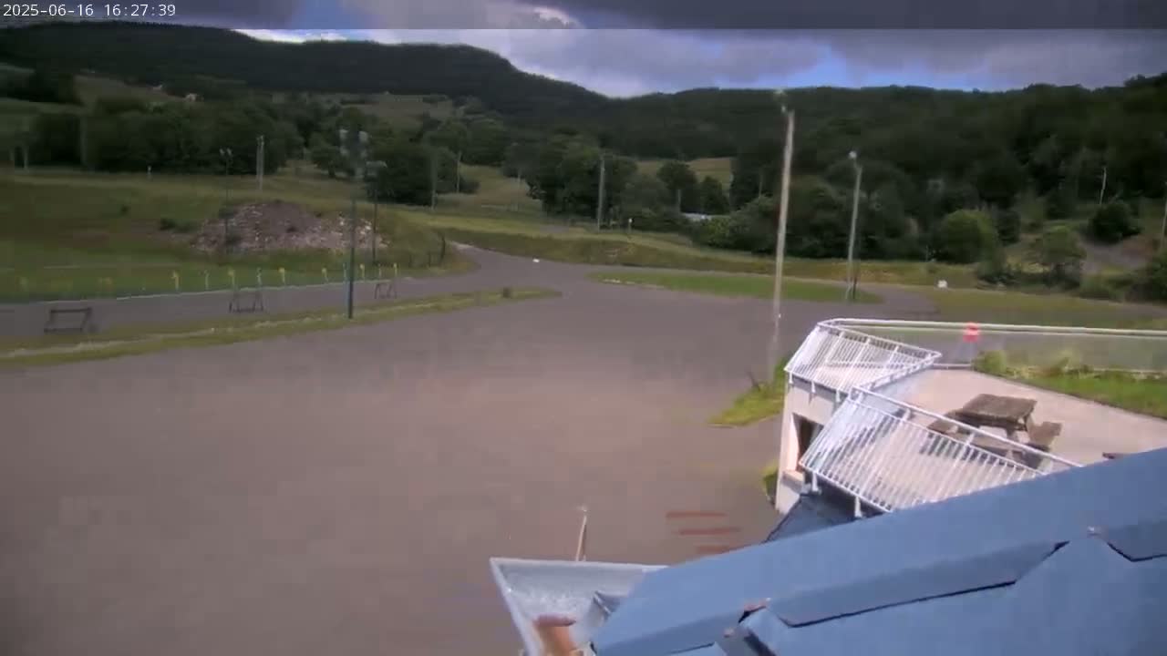 A mostly cloudy sky overlooks an asphalt area with a building featuring a patio, picnic tables, and a water slide, all set against a backdrop of green hills.