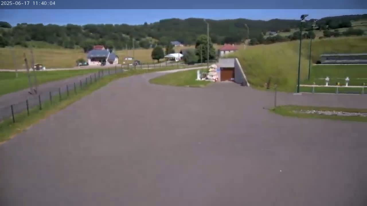 A paved road curves through a rural area on a sunny day, passing by buildings and a grassy hillside with distant trees.