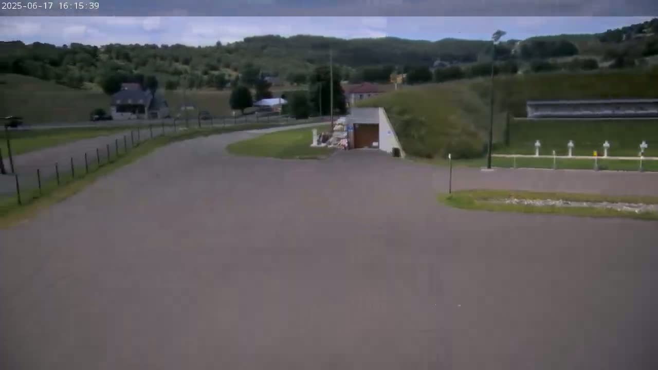 A paved area curves around a small building with a pile of debris nearby, under a partly cloudy sky, with a hilly, green landscape in the background.