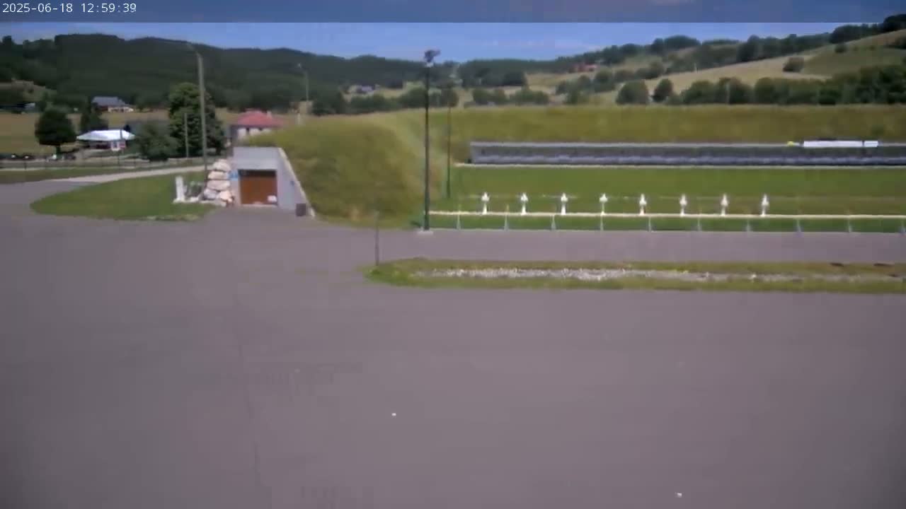 A paved area curves toward a small building near a shooting range on a sunny day with rolling green hills in the background.