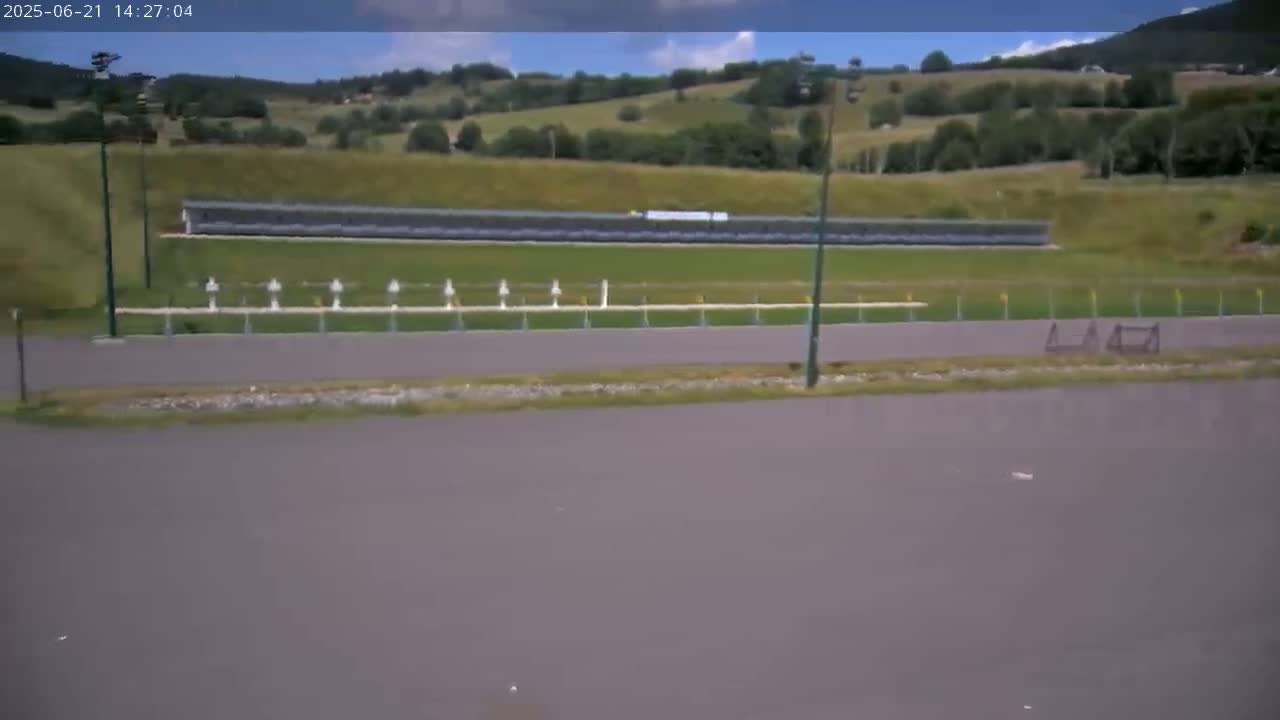 A shooting range with a long row of targets is situated on a paved area, overlooking a grassy hillside under a partly sunny sky.