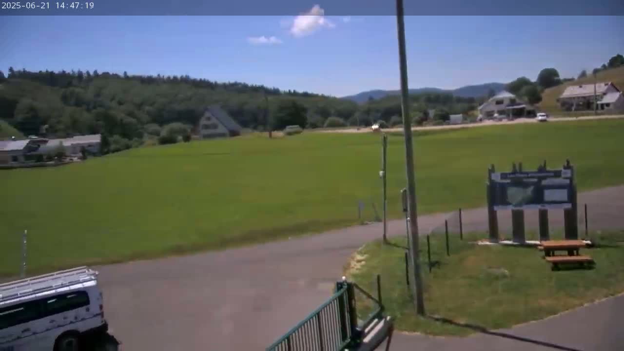 A large grassy field, under a mostly sunny sky, is situated between a road and a line of trees and buildings.