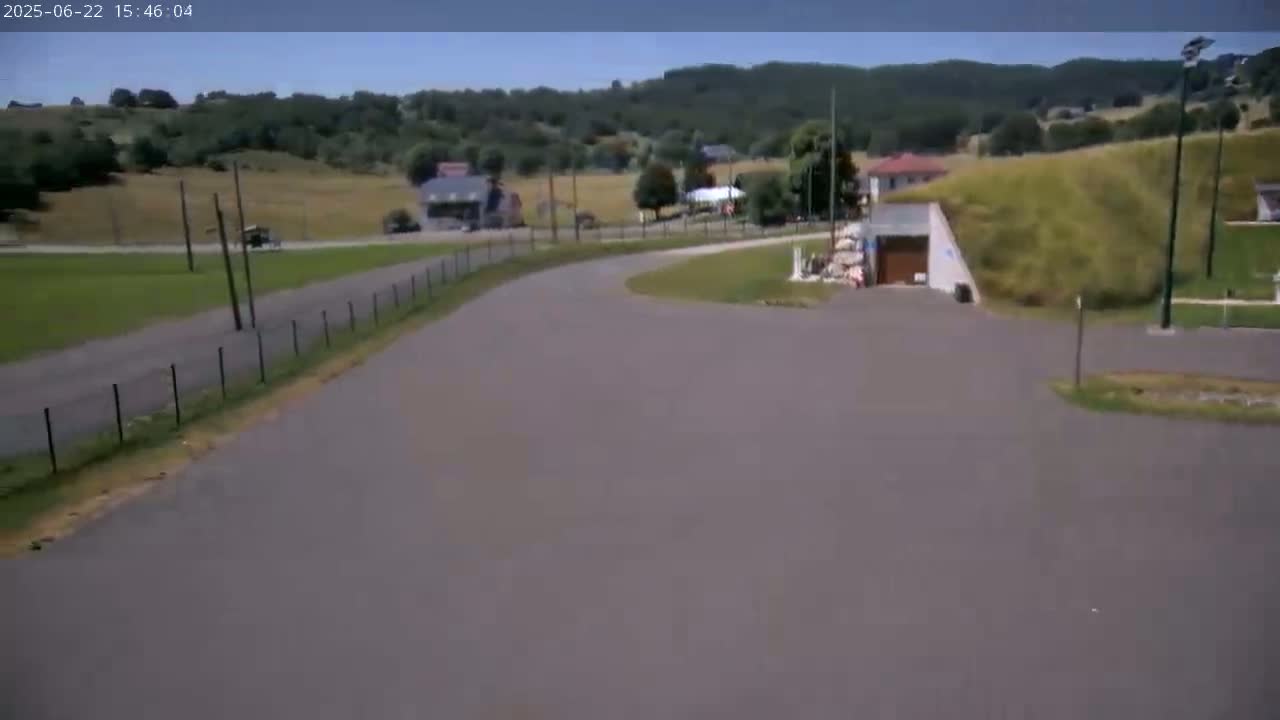 A paved road curves through a rural landscape on a sunny day, with hills and a few buildings in the distance.