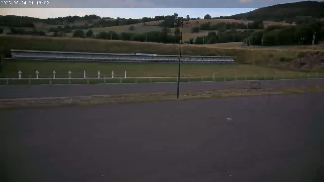 A long, covered structure sits on a grassy field next to a paved track, all under an overcast sky.