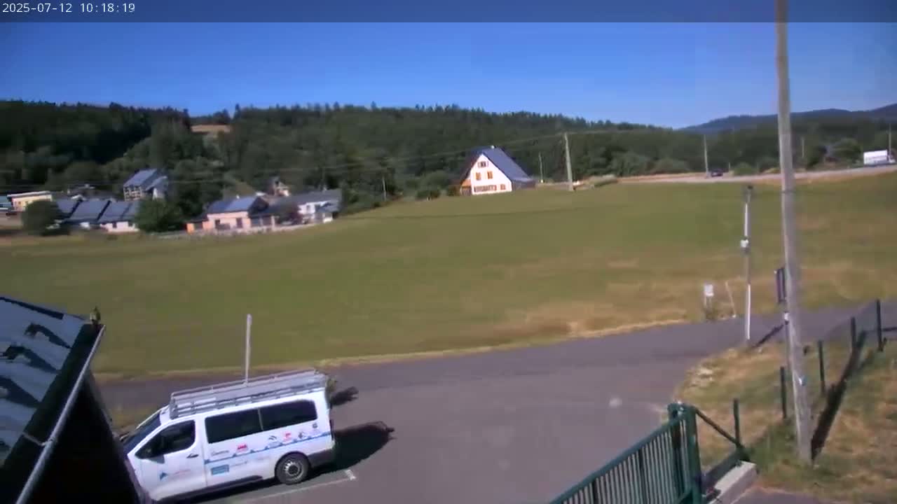 A white van is parked in a lot next to a grassy field, with a cluster of houses and a forest visible in the background under a clear, sunny sky.
