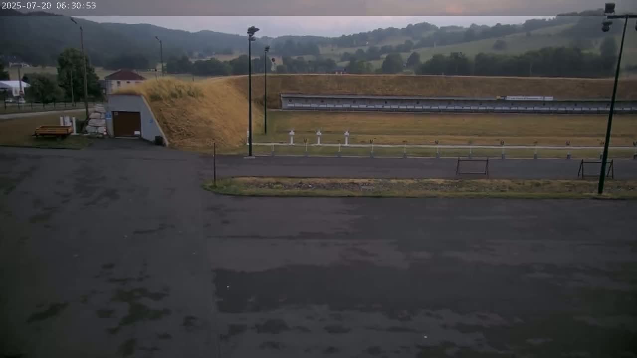 An outdoor shooting range with a covered structure and a small building is visible under an overcast sky.