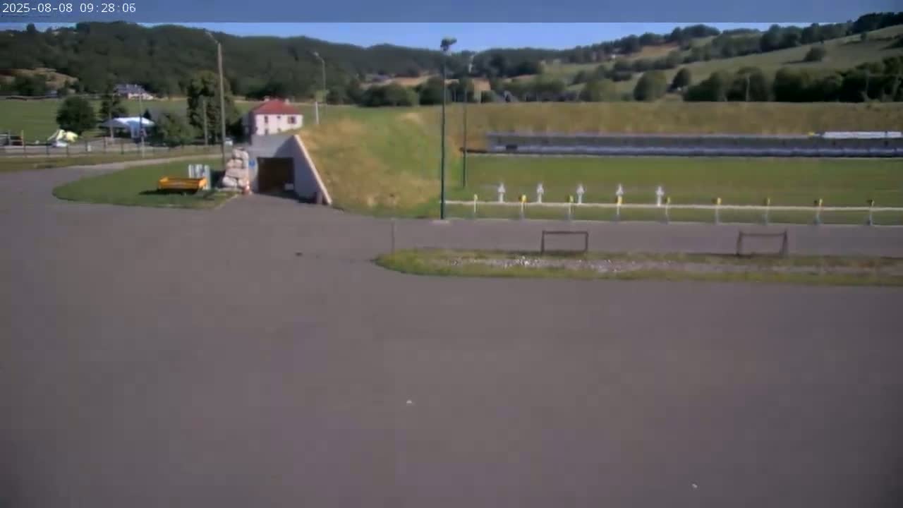 A paved area with a grassy embankment leading to a building,  a shooting range in the background, all under a sunny sky.