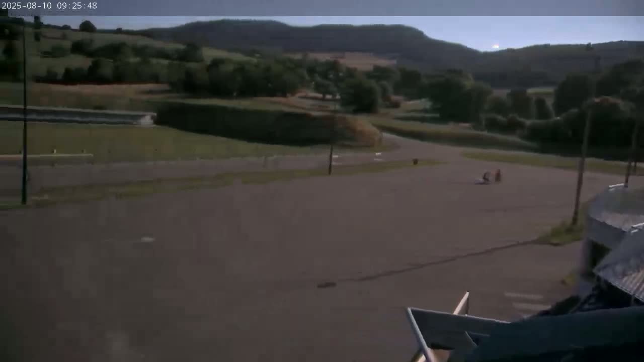 Two people are on a paved area near some green hills under an overcast sky.