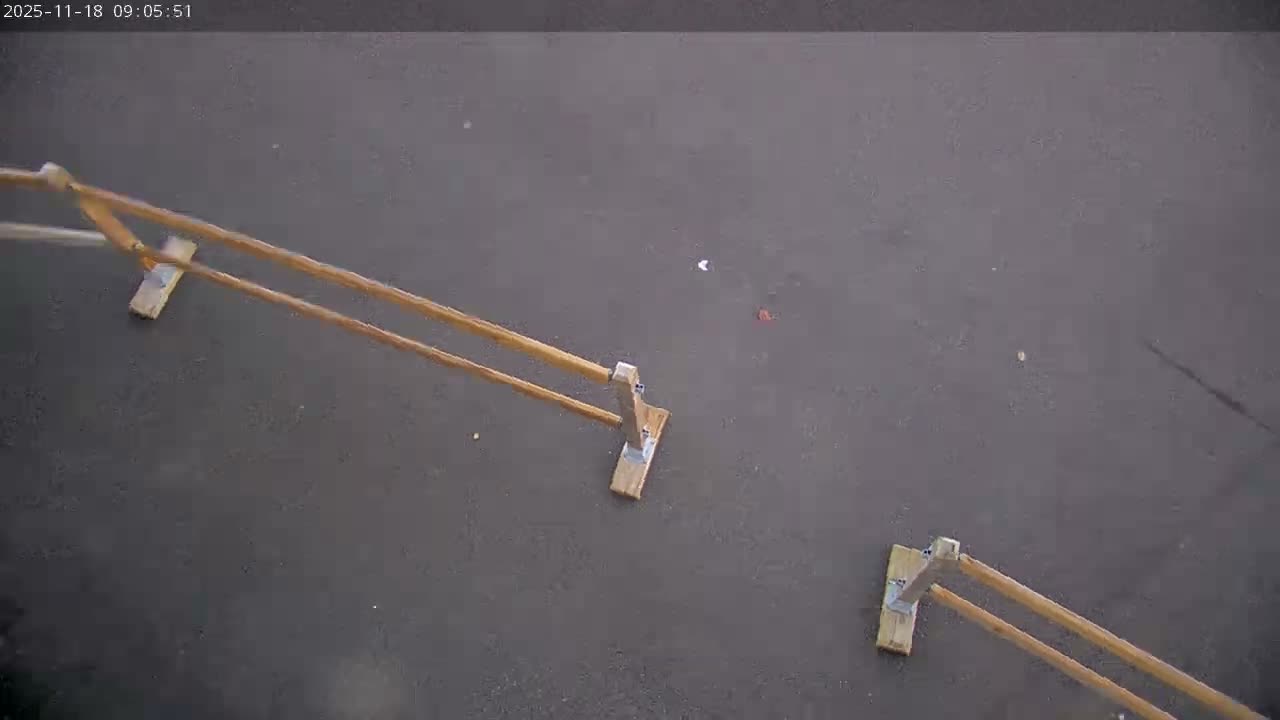 An overhead view shows a dry, dark grey paved surface with three wooden structures, each featuring a flat base and two parallel angled poles, along with scattered small bits of debris, all under an overcast sky.