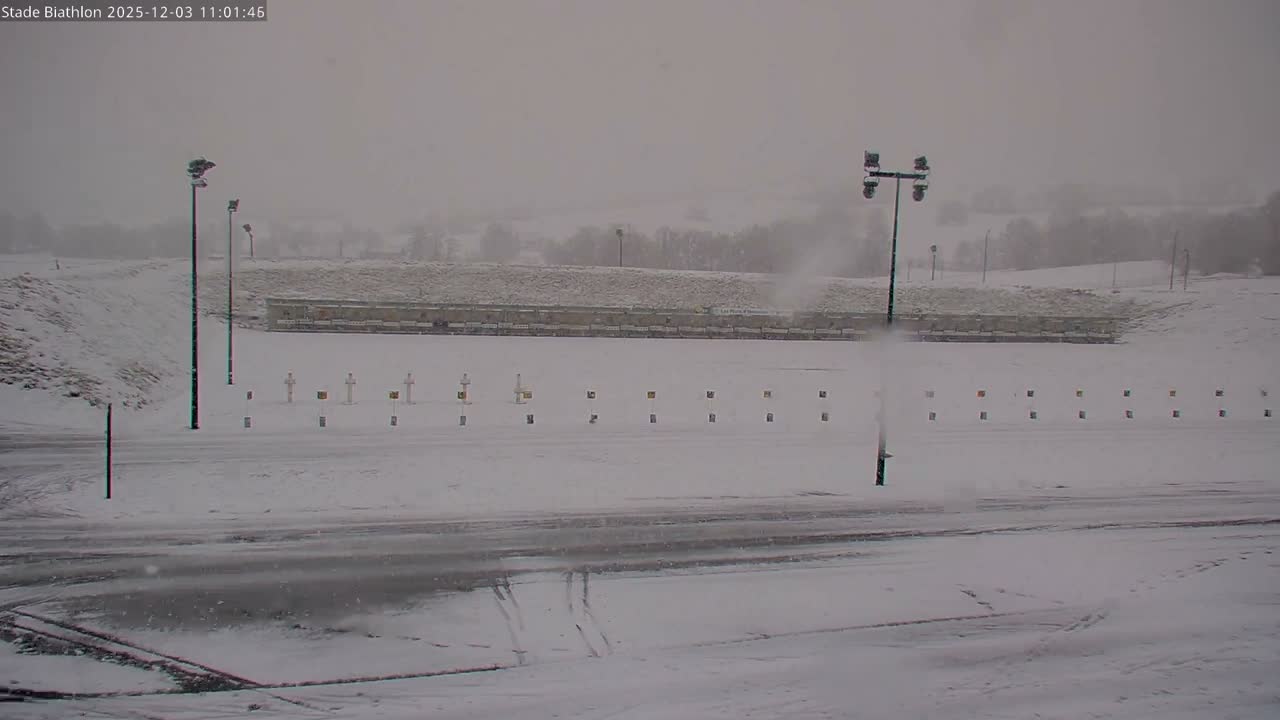 A snow-covered outdoor sports area, featuring rows of white cross-shaped targets and square markers, along with tall floodlight poles, lies under a uniformly gray, overcast sky with light snow actively falling.