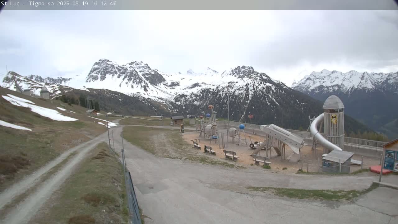 A playground sits in a mountainous area with snow-capped peaks under an overcast sky.