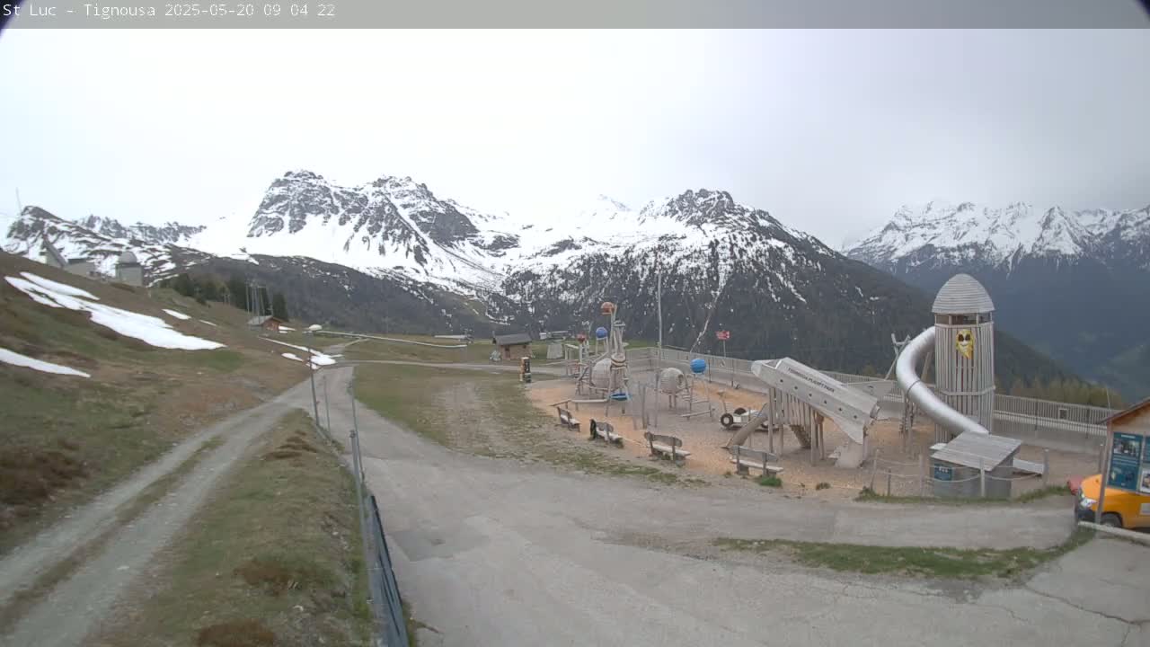 A playground sits in a mountain valley under an overcast sky, with snow-capped mountains in the background.