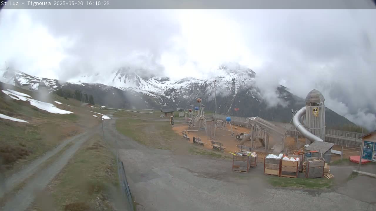A playground with a large slide sits below a mountain range partially obscured by clouds, with patches of snow visible on the ground and mountains.