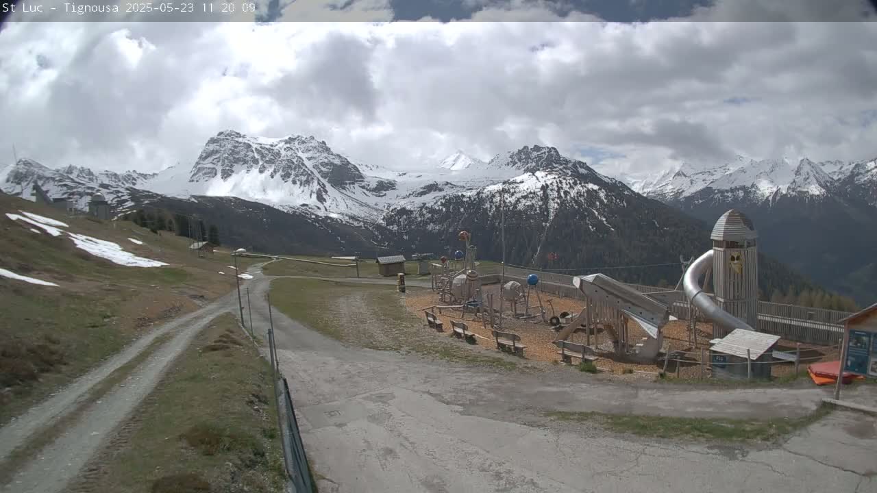 A playground with a large metal slide sits in a mountainous area with patchy snow under a partly cloudy sky.