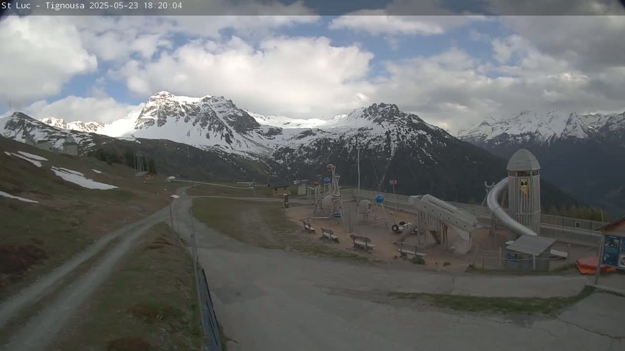 A playground sits near a roadway overlooking snow-capped mountains under a partly cloudy sky.