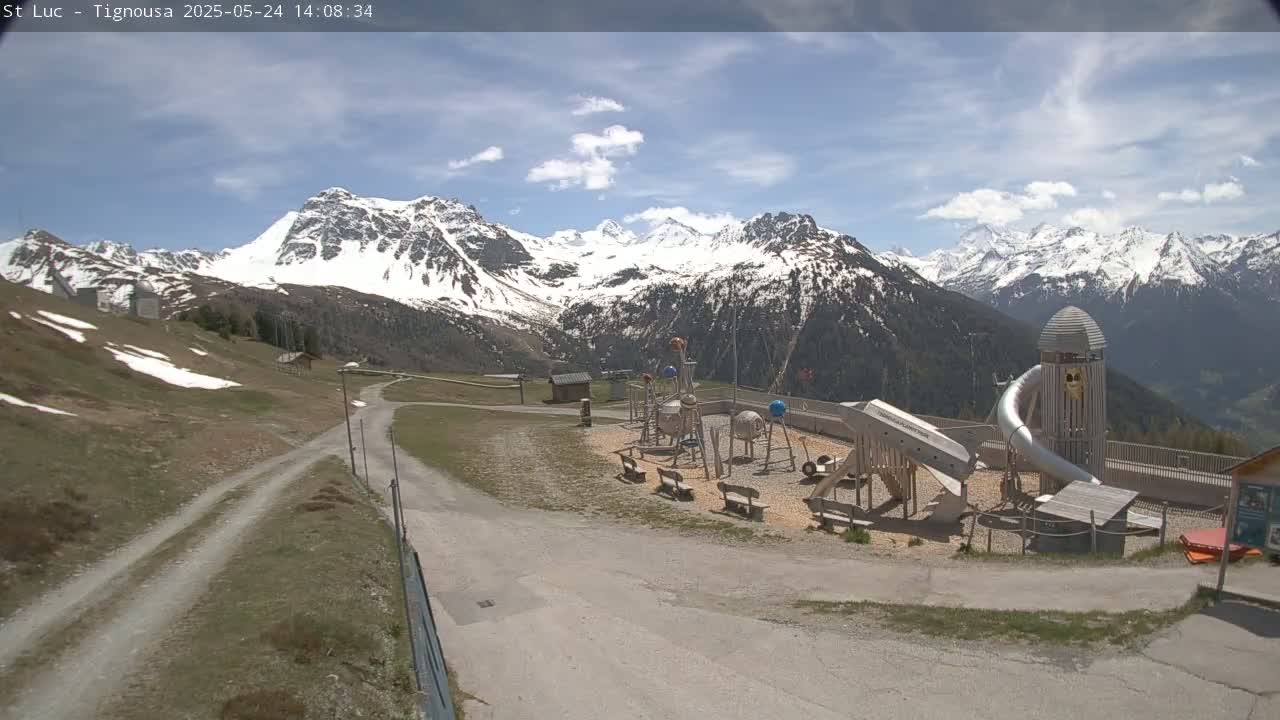 A playground sits near a dirt road in front of a snow-capped mountain range under a partly cloudy sky.