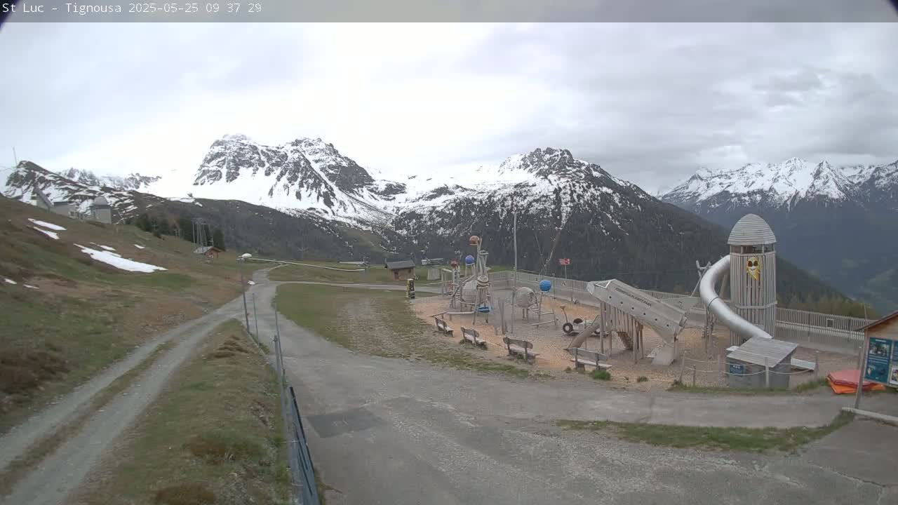 A playground with a large slide sits in front of snow-capped mountains under an overcast sky.