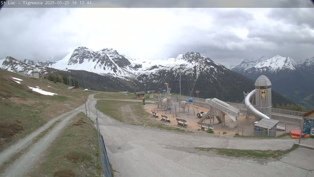 A playground sits on a gravel lot overlooking snow-capped mountains under a cloudy sky.