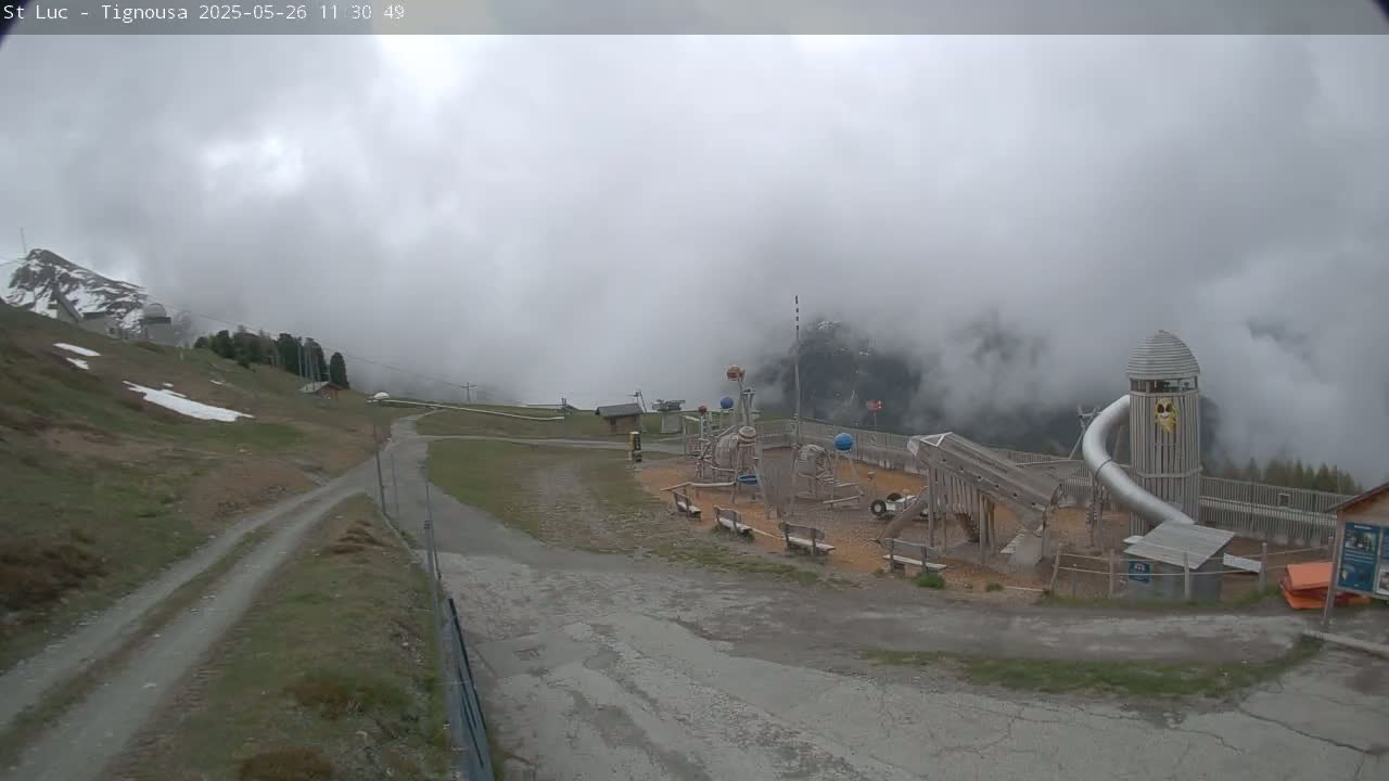 A playground with a large metal slide is situated on a mountainside shrouded in fog, with a gravel path leading to it.