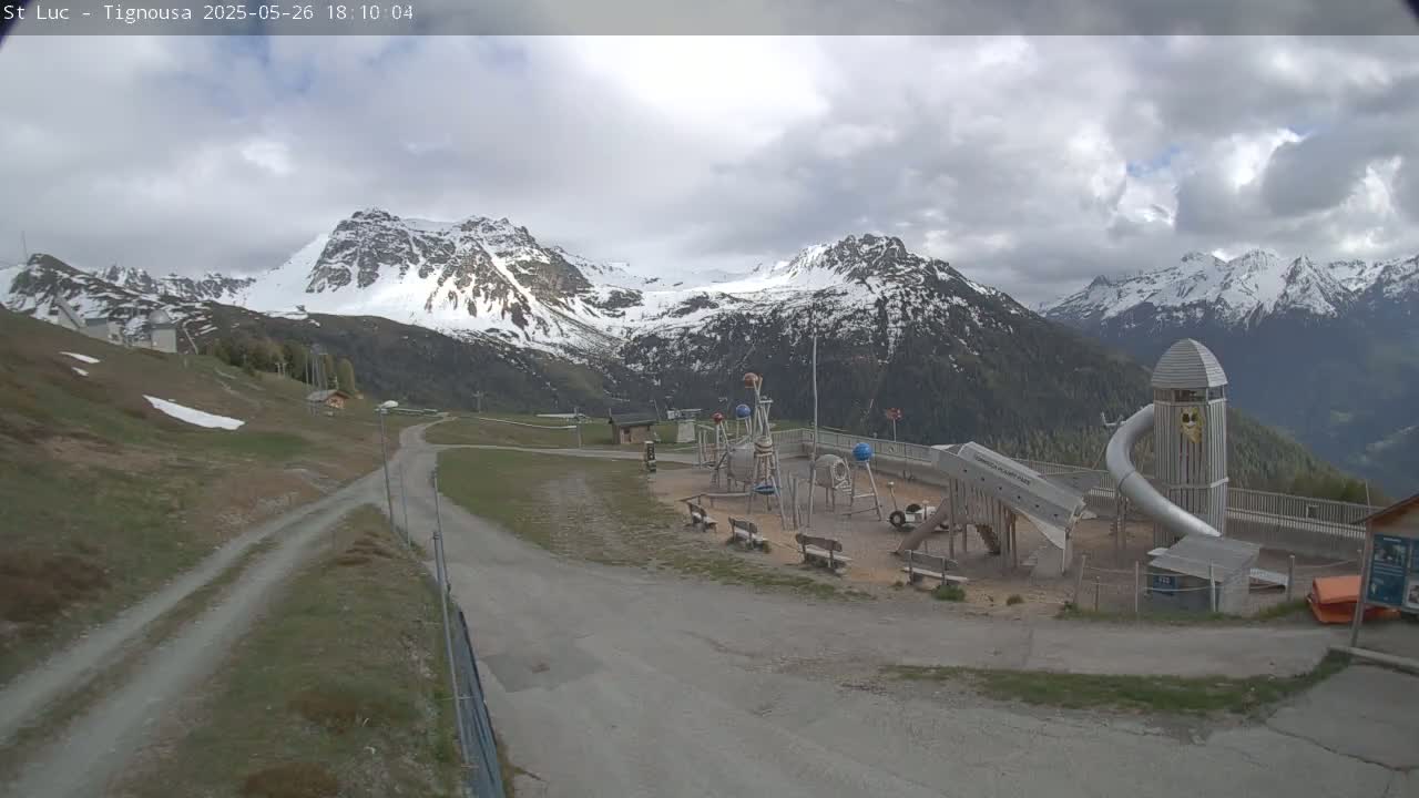 A playground with a large metal slide sits below snow-capped mountains under a cloudy sky.