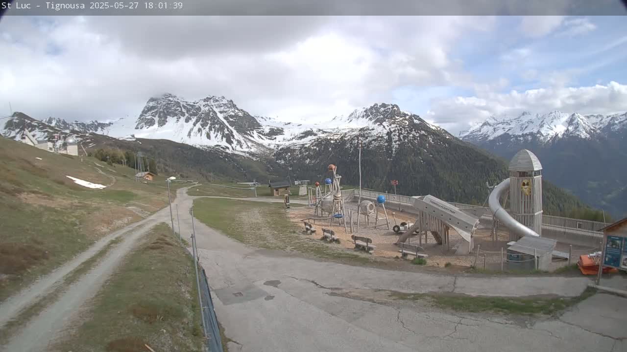 A playground with a long slide sits near a paved road, overlooking snow-capped mountains under a partly cloudy sky.