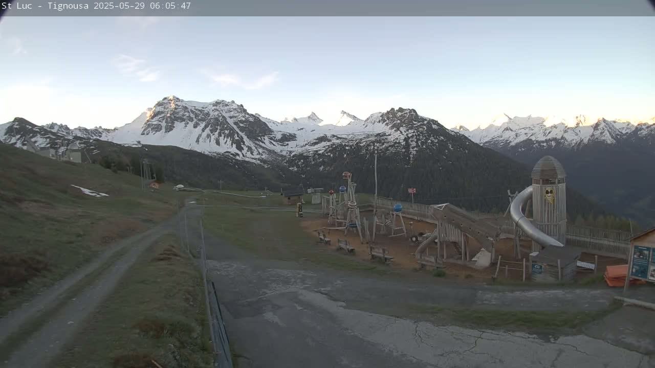 A playground sits near a gravel road with snow-capped mountains in the background under a clear sky.