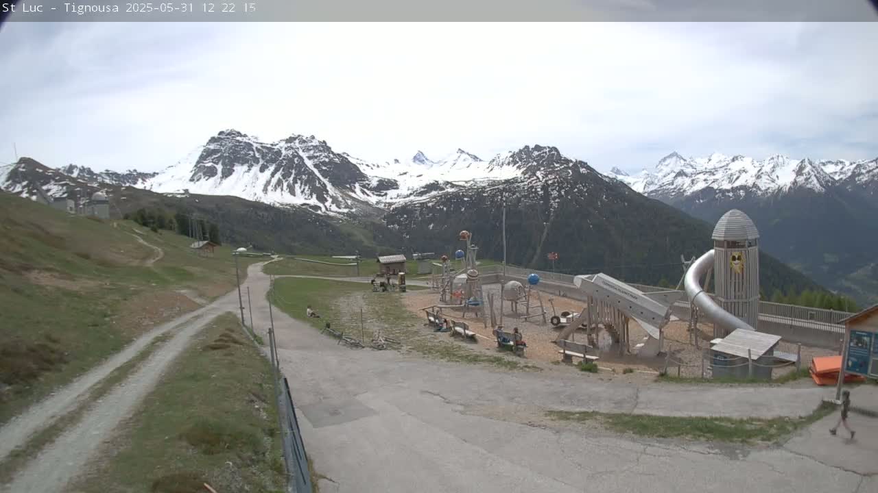 A playground with a large metal slide sits below a mountain range partially covered in snow under an overcast sky.