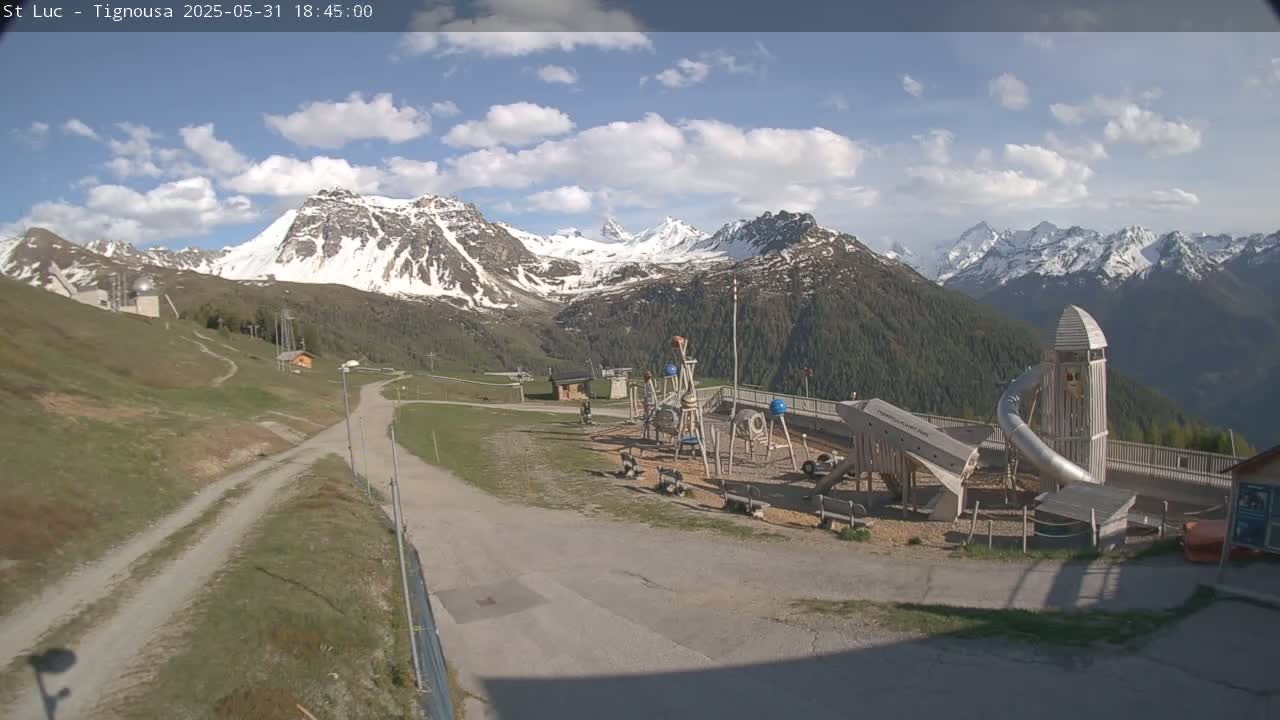 A playground with a large slide sits in a grassy area overlooking snow-capped mountains under a partly cloudy sky.