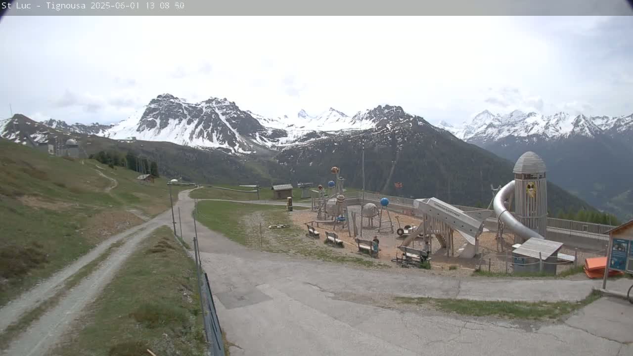 A playground with a large slide is situated on a hill overlooking snow-capped mountains under an overcast sky.