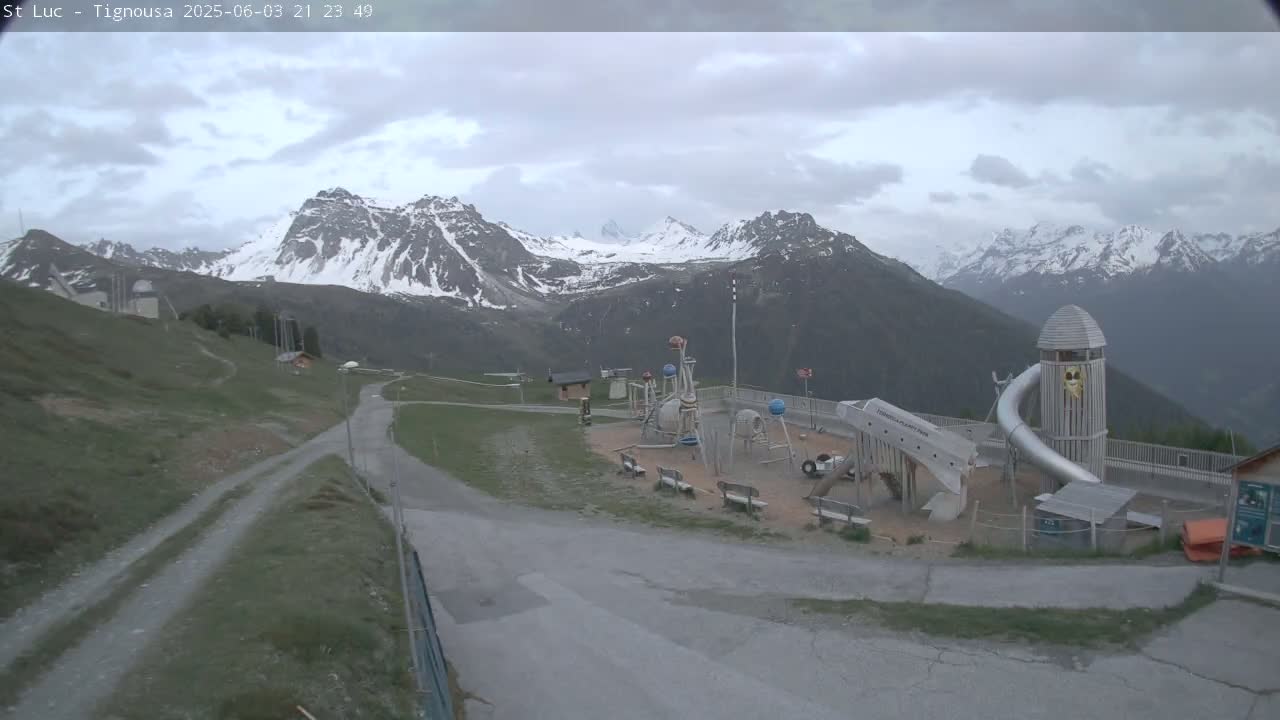 A playground sits in a mountainous area under a cloudy sky, with snow-capped peaks in the background.