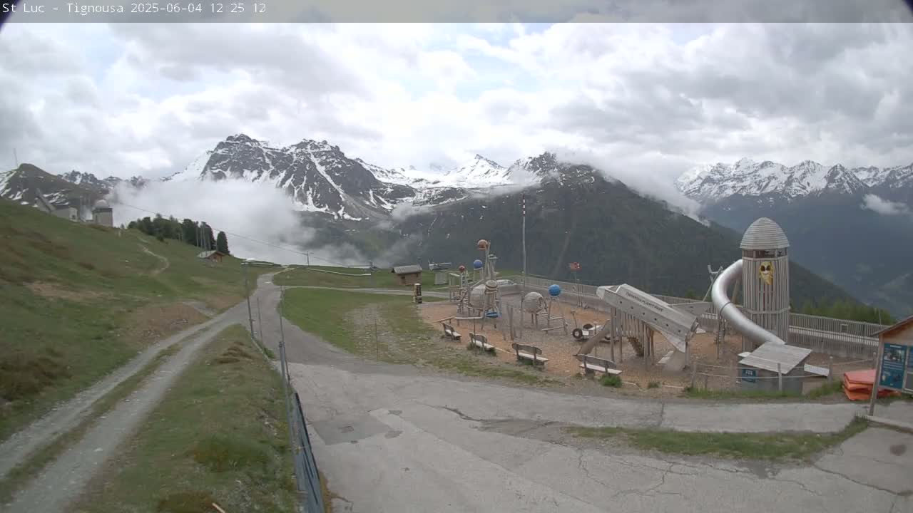 A playground sits on a mountainside overlooking snow-capped mountains partially obscured by clouds.