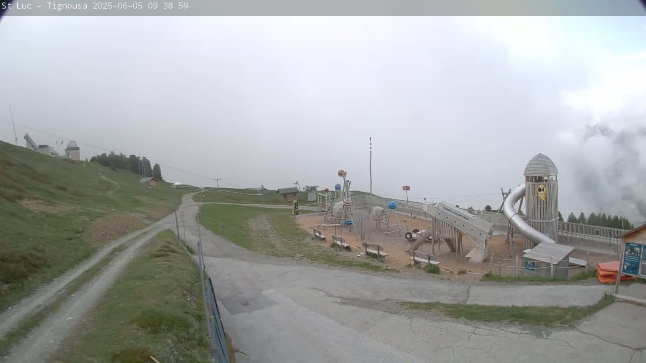A playground with a large metal slide sits on a grassy hilltop in foggy conditions, with a dirt road leading to it.