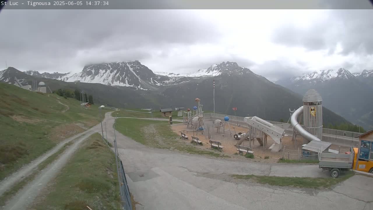 A playground with a large metal slide sits below snow-capped mountains under an overcast sky.