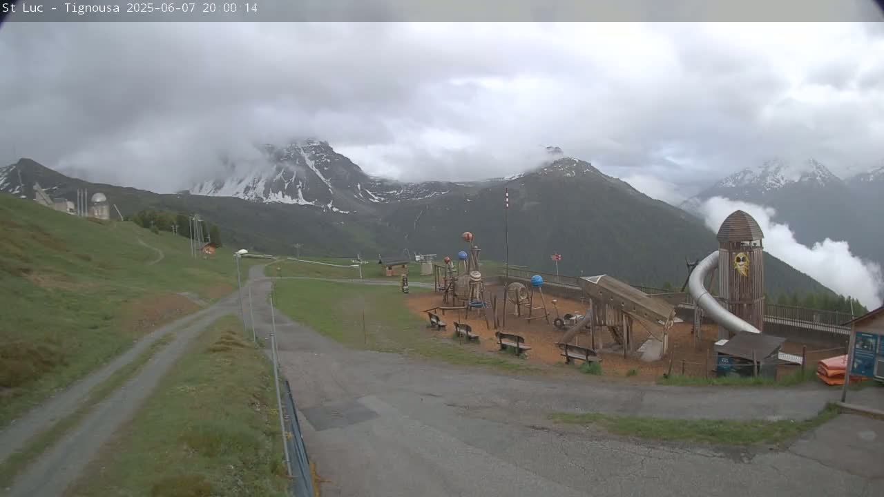 A playground sits in a grassy area near a paved road, with overcast skies and snow-capped mountains in the background.
