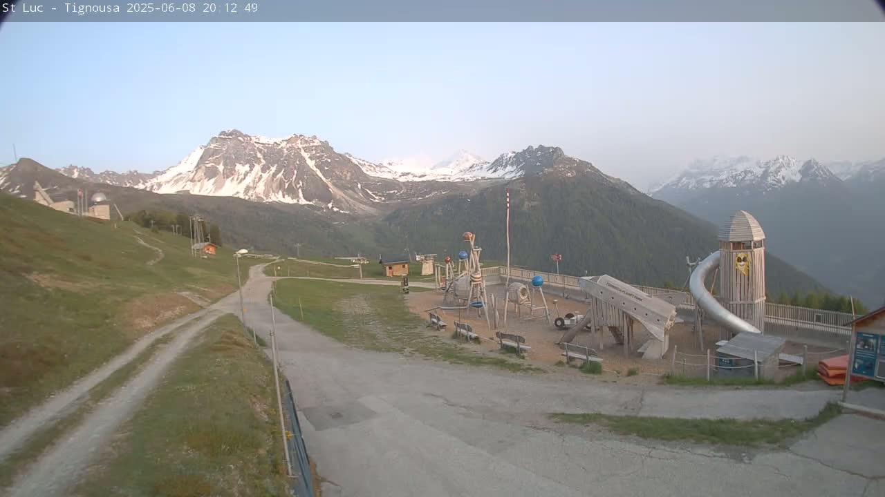 A playground sits near a mountain road, with snow-capped mountains in the background under a clear sky.