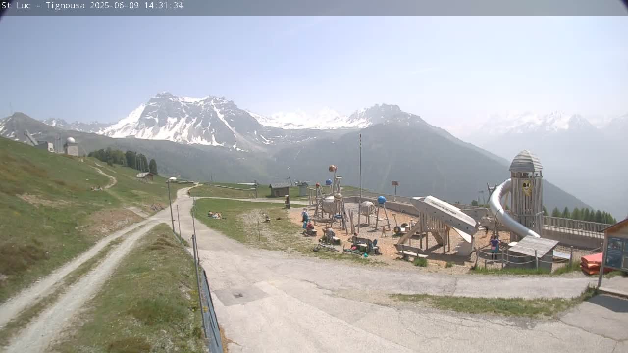 A playground with a large slide is situated on a hill overlooking snow-capped mountains under a hazy, sunny sky.