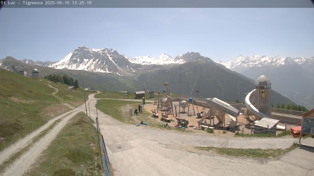 A playground sits on a grassy hill overlooking snow-capped mountains under a clear, sunny sky.
