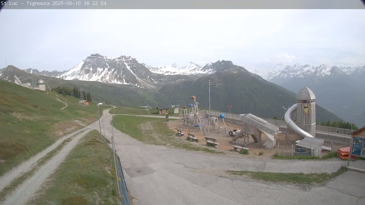 A playground with a large metal slide is situated on a hill overlooking snow-capped mountains under an overcast sky.