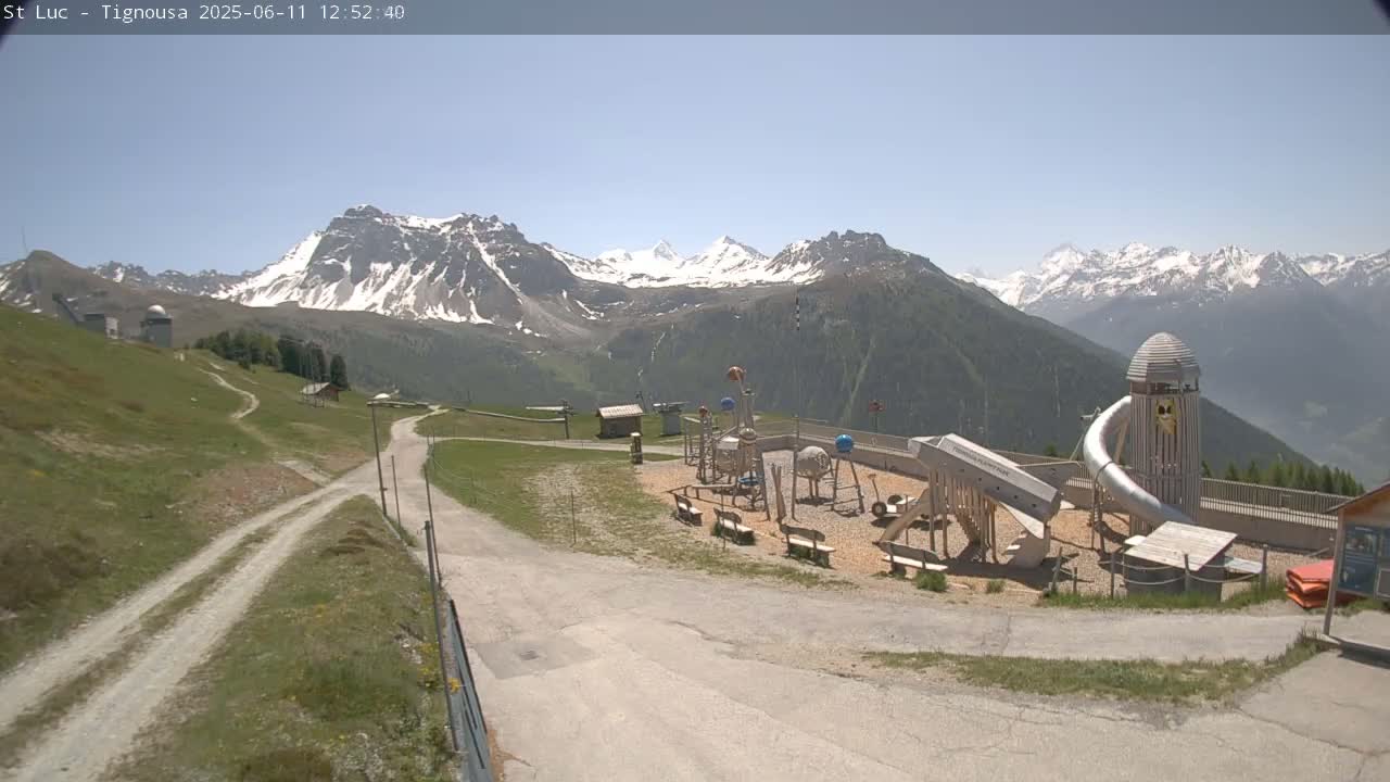 A playground sits near a mountain road overlooking snow-capped mountains under a clear, sunny sky.