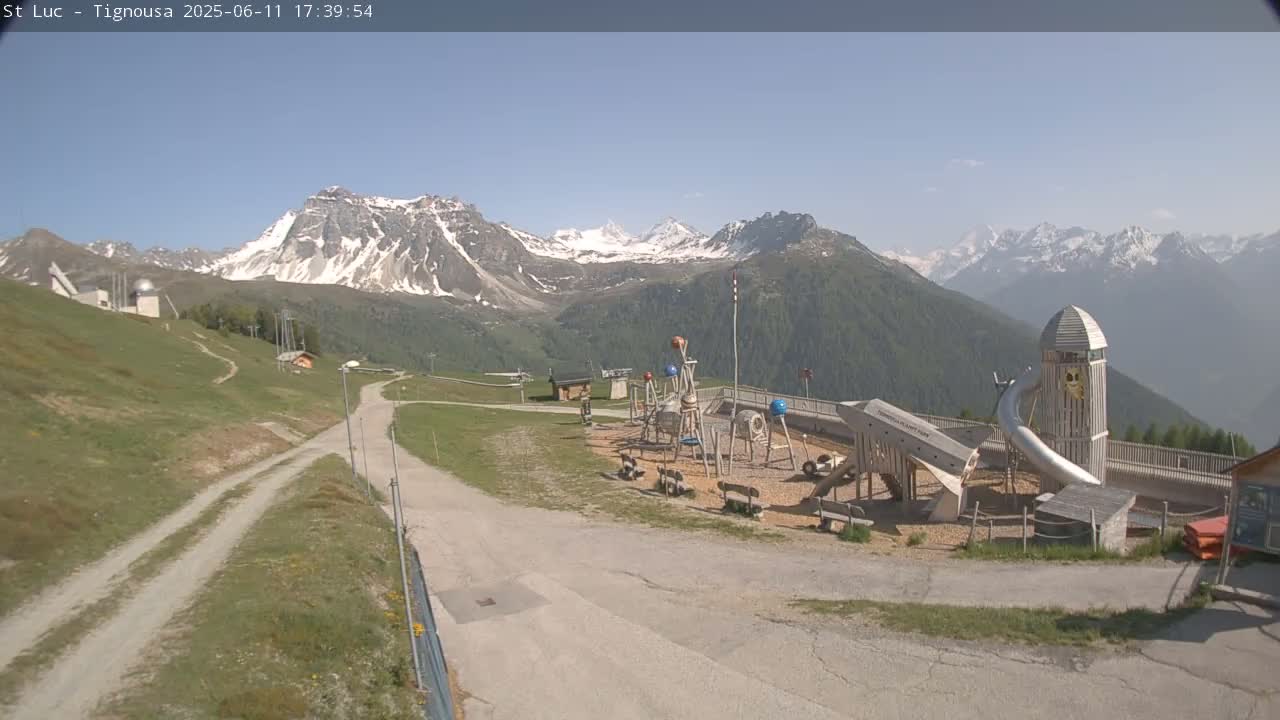 A playground sits on a grassy hill overlooking snow-capped mountains under a clear, sunny sky.