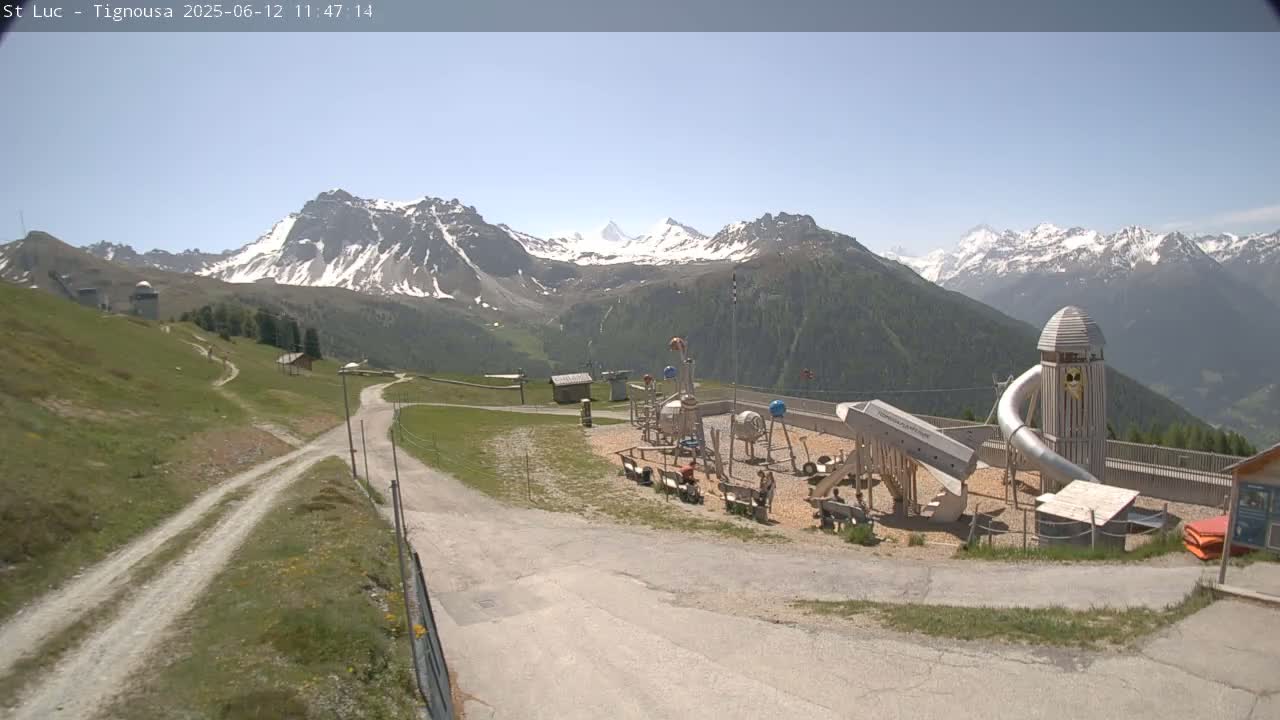 A playground sits in a mountainous area with snow-capped peaks visible in the background under a clear, sunny sky.