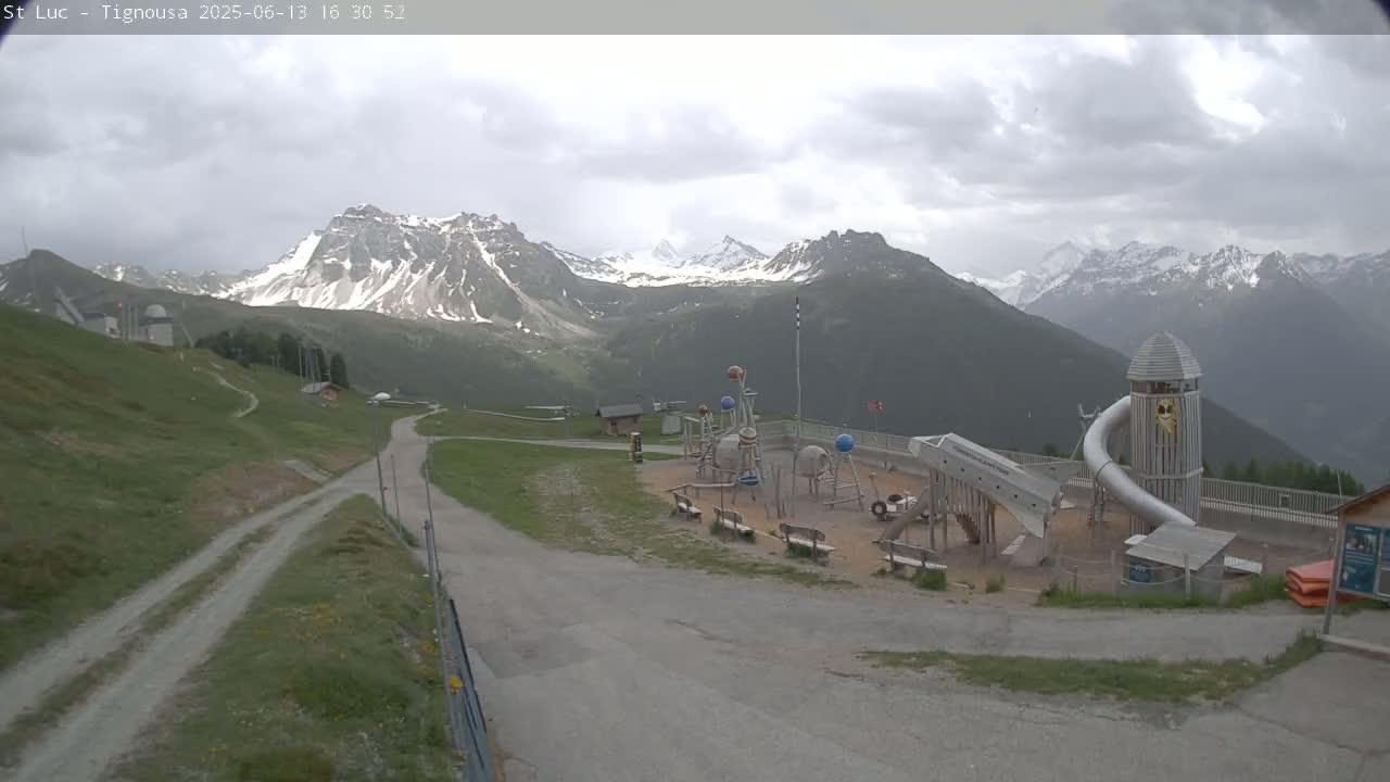 A playground with a large slide is situated on a grassy area overlooking a mountain range under a cloudy sky.